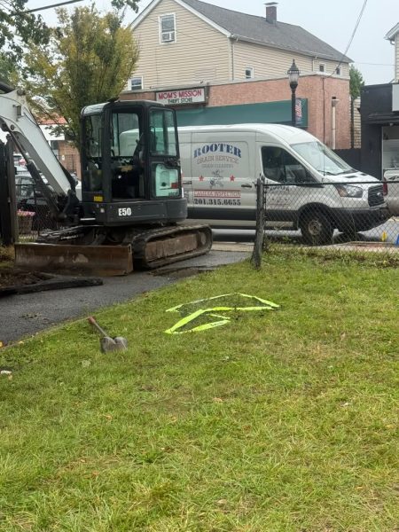 A mini excavator and the Rooter Drain Service van on a front lawn, with utility marking paint on the grass.