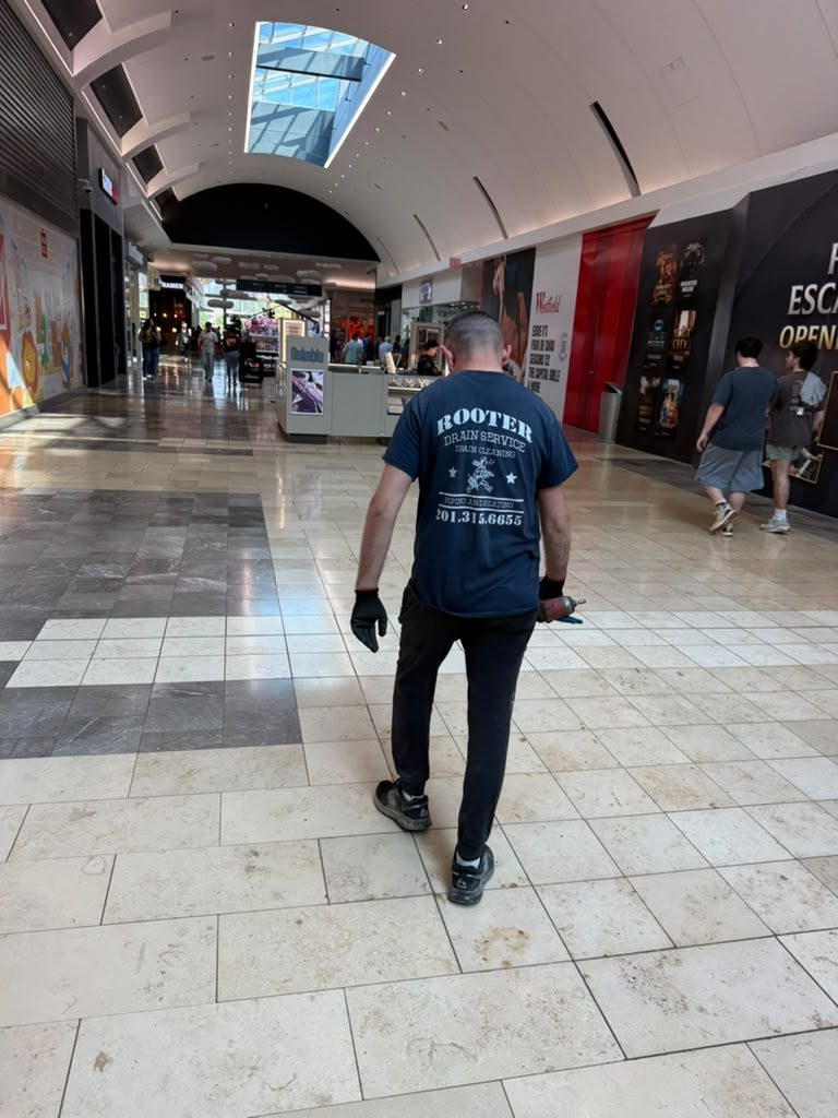 A technician in a Rooter Drain Service shirt walking through a tiled retail arcade carrying tools.
