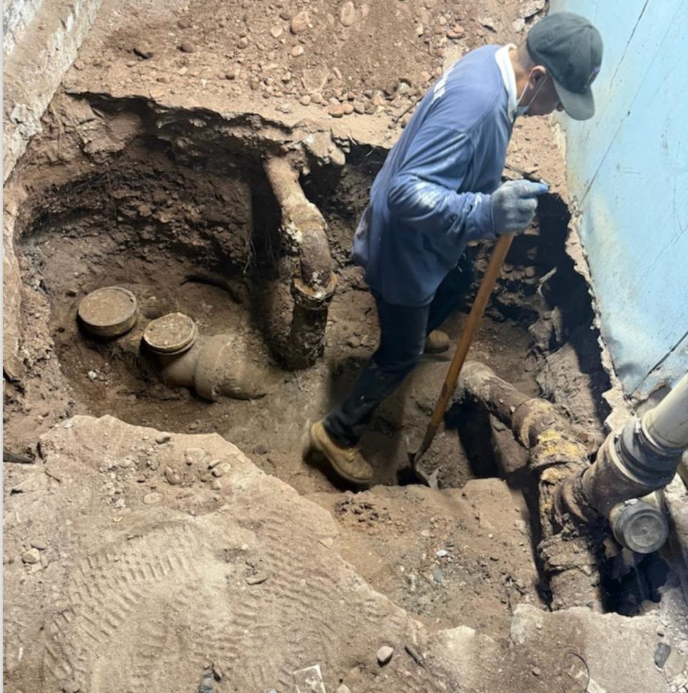 A technician standing in an open excavation, working on a pipe at the bottom of the hole.